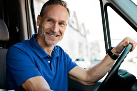 Photograph of the interior of a truck or van with a man smiling towards the camera resting his hand on the wheel